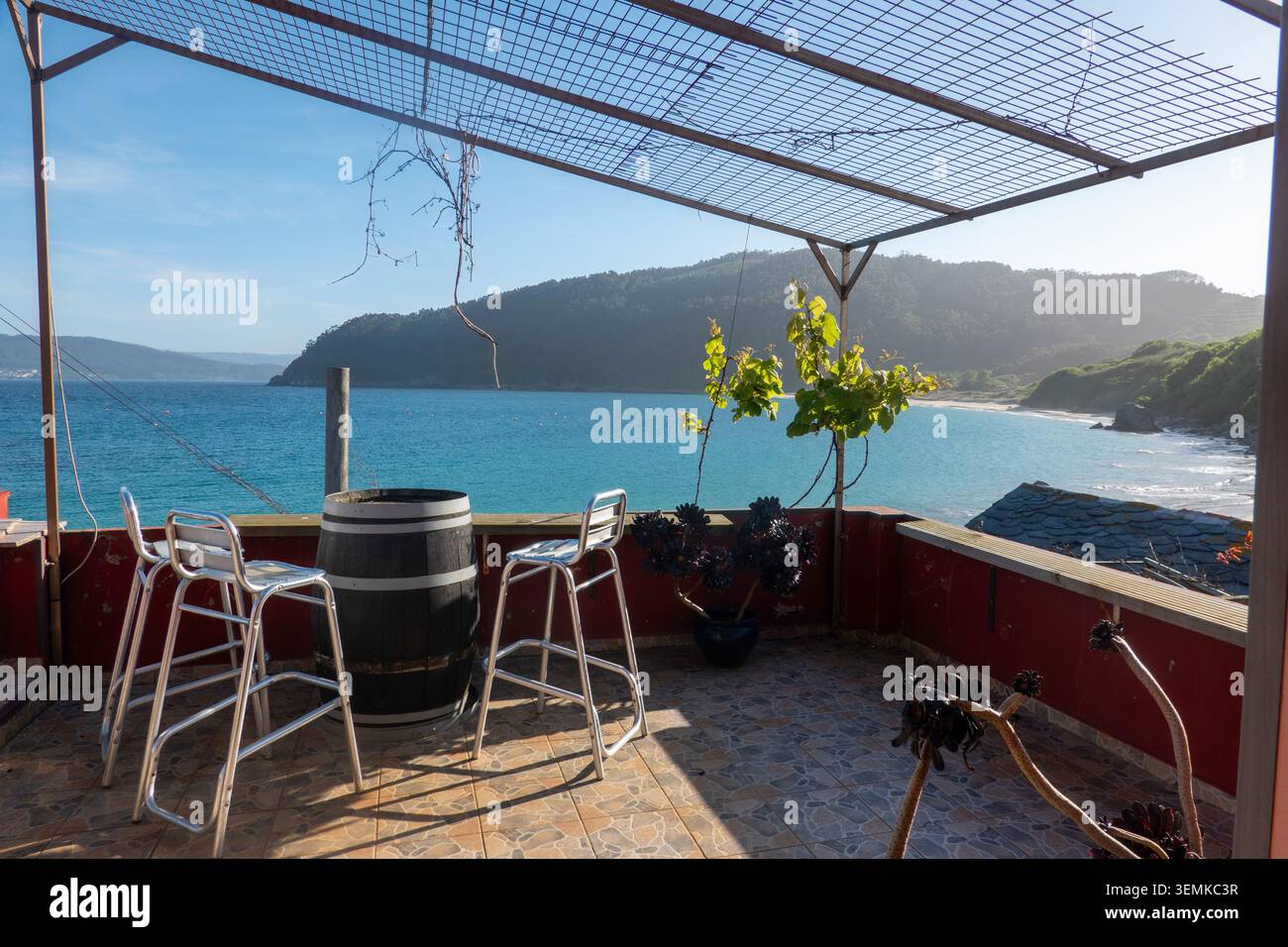 Une terrasse ensoleillée au port de Bares avec une table tonneau et des tabourets métalliques, offrant une vue imprenable sur la mer turquoise, la plage et les montagnes verdoyantes. Banque D'Images