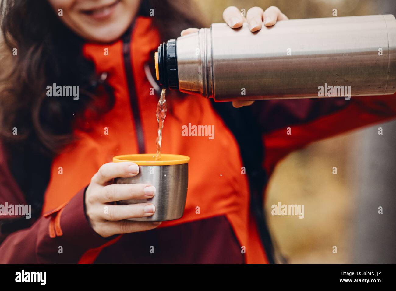 Femme souriante verse la boisson chaude du thermos en métal dans la tasse à l'extérieur. loisirs forestiers saisonniers, tenue de camping fonctionnelle, trekking par temps froid Banque D'Images