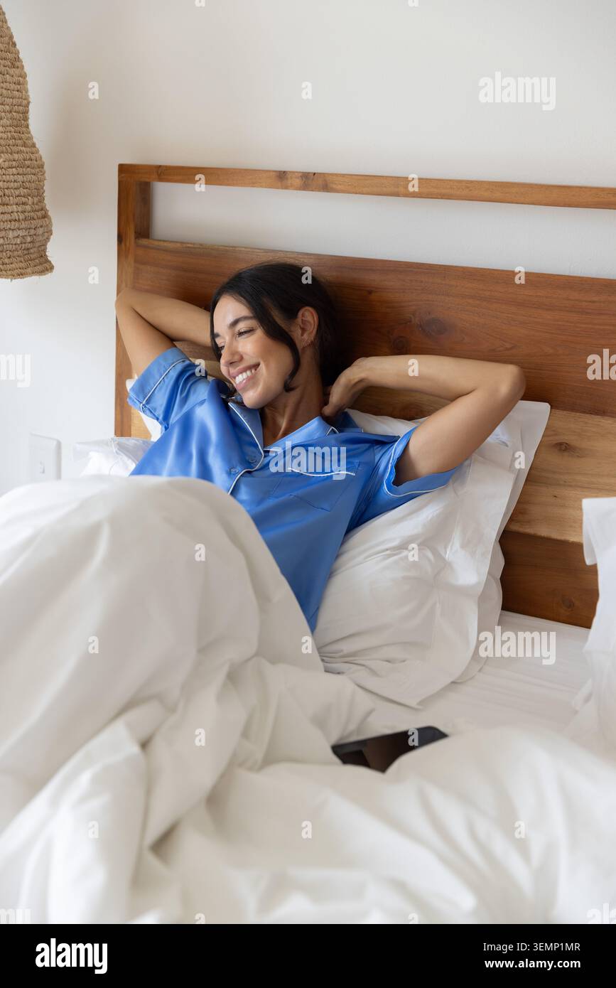 Femme en pyjama en satin haut étirant les bras derrière la tête dans le lit avec literie blanche et smartphone Banque D'Images