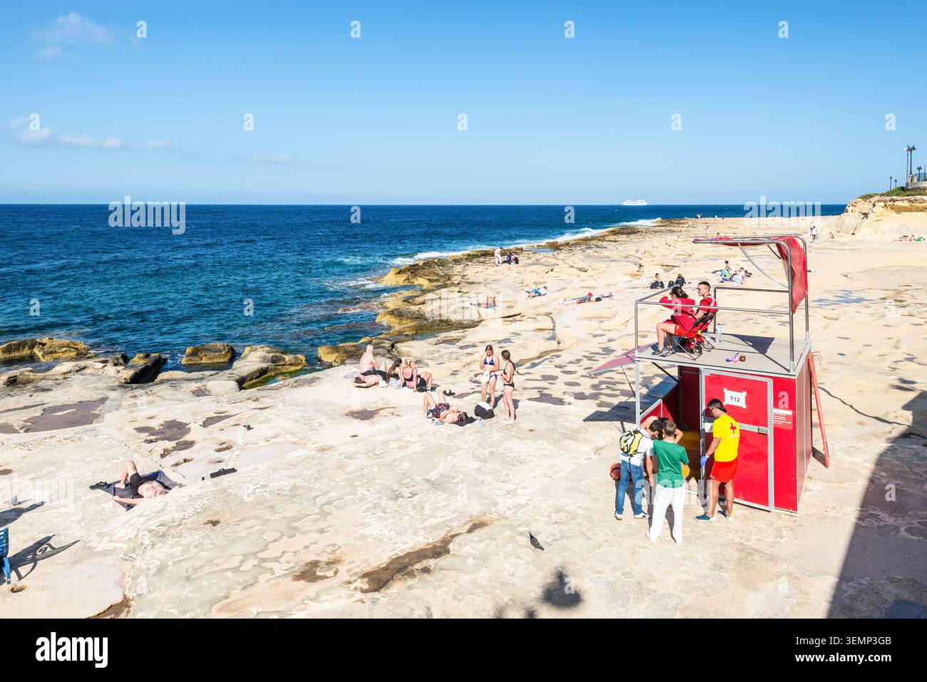 Sliema, Malte – 16 mai 2025 : des sauveteurs surveillent les nageurs et les bains de soleil depuis une tour rouge le long de la côte rocheuse de Sliema Beach sous un soleil éclatant Banque D'Images
