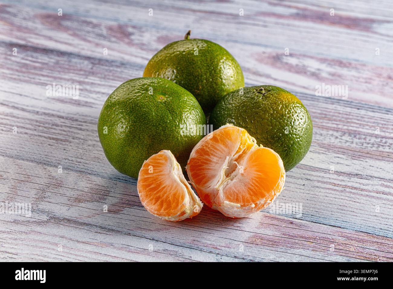 Trois mandarines vertes non mûres à la peau texturée reposent sur une surface en bois altérée à côté d'un demi-fruit pelé révélant un intérieur orange juteux Banque D'Images