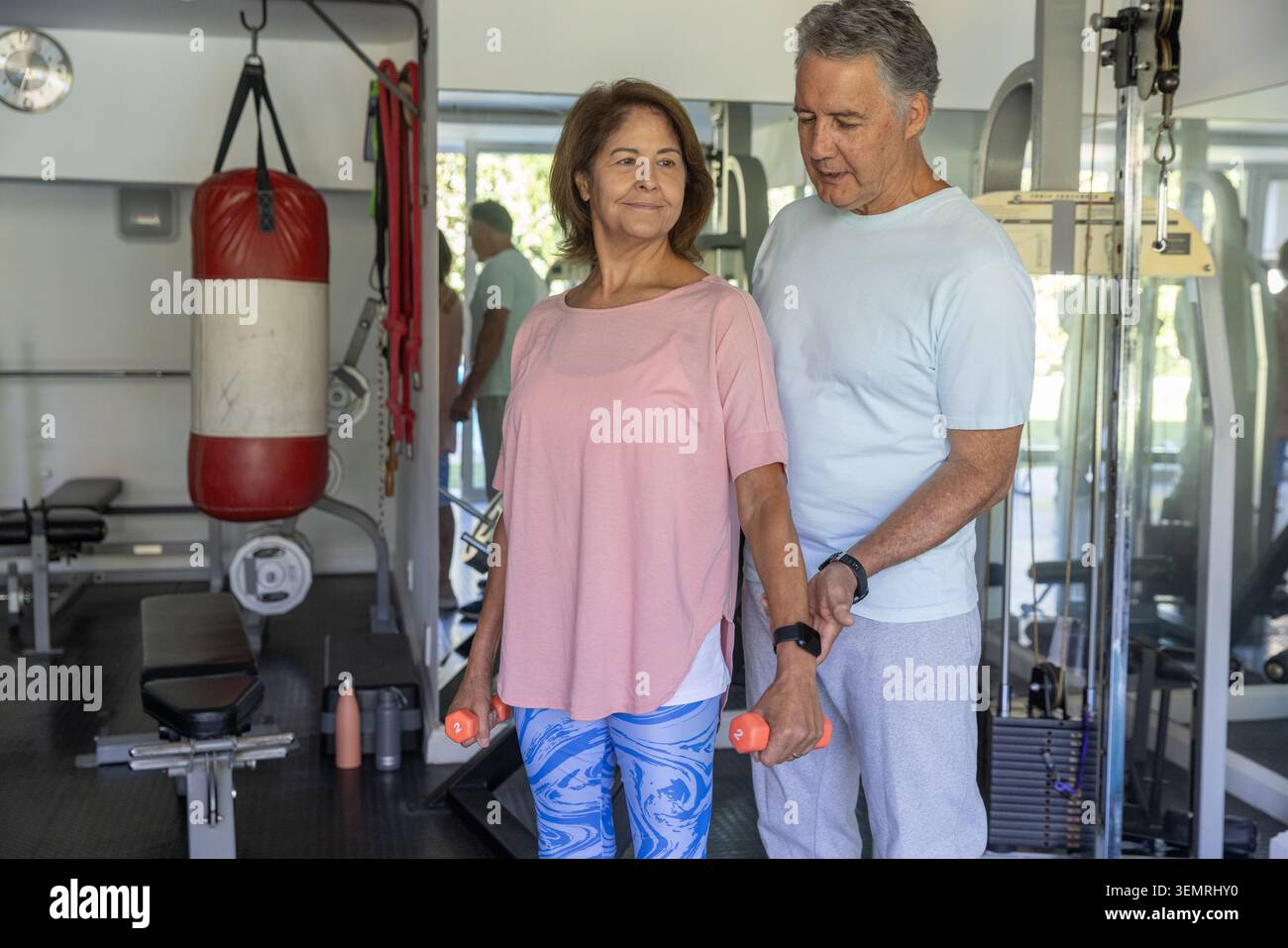 Couple : homme mûr et femme âgée exerçant dans la salle de gym, femme en haut rose tenant des haltères orange Banque D'Images