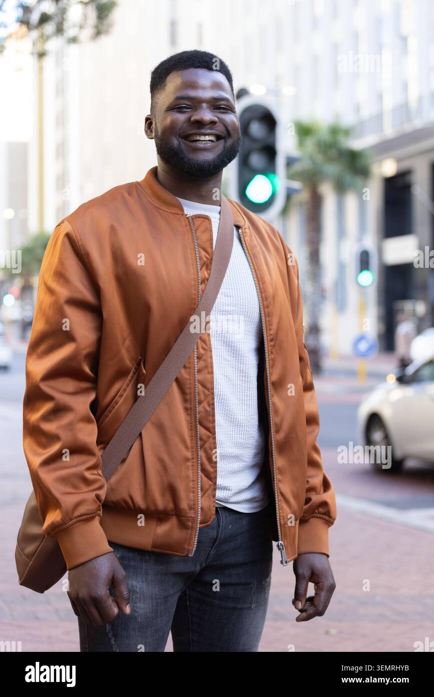 Homme afro-américain souriant, debout sur le trottoir portant blouson bomber, sangle de sac, lumière verte Banque D'Images