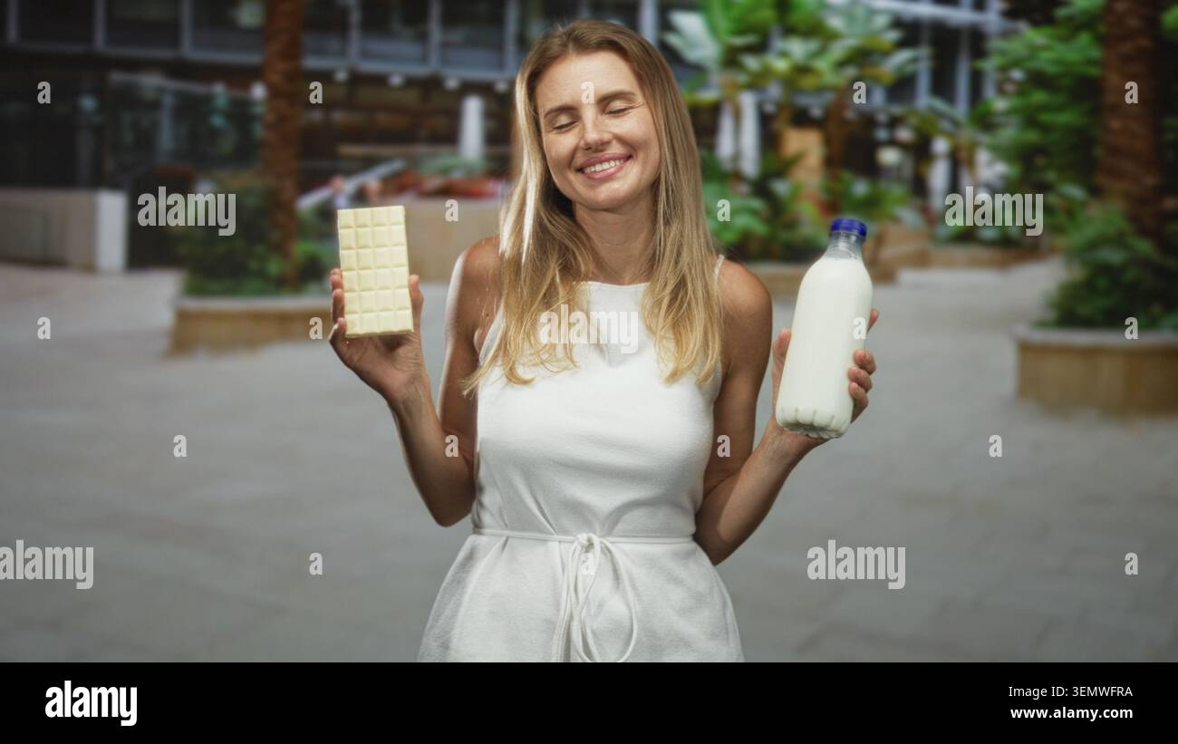 Femme tient une barre de chocolat blanc et une bouteille de lait avec les yeux fermés et souriant dans l'atrium du bâtiment ; plaisir de contentement. Banque D'Images