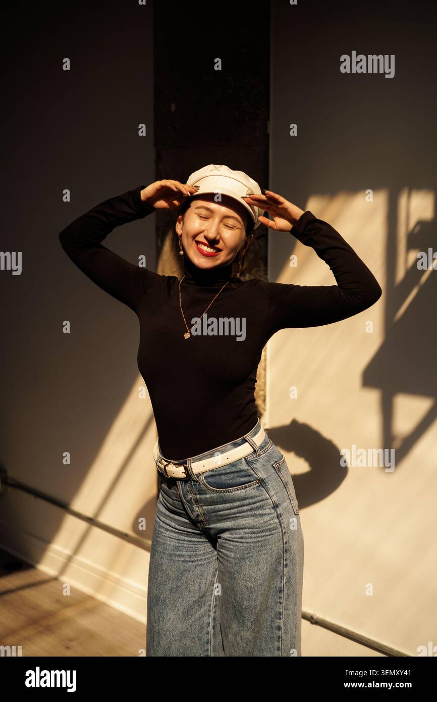 Une femme se tient dans une pièce avec une lumière vive et des ombres. Elle porte une casquette et une chemise noire à manches longues. Ses jeans sont larges et elle sourit en posant Banque D'Images