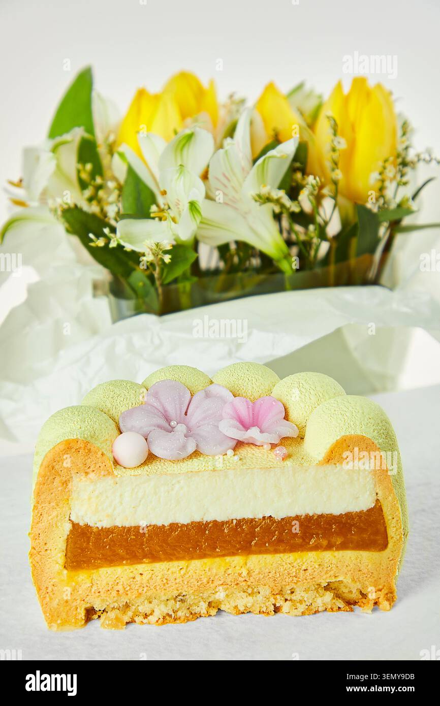 Coupe transversale de gâteau de mousse jaune montrant mousse crémeuse et garniture d'abricot avec bouquet de printemps flou en arrière-plan. Dessert festif élégant. Banque D'Images