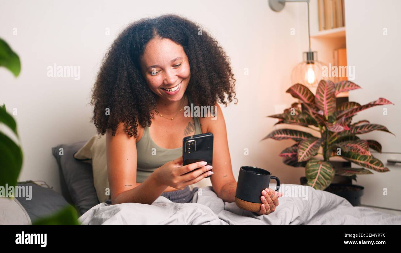 Femme souriante se relaxant dans le lit avec smartphone et café dans une chambre confortable Banque D'Images