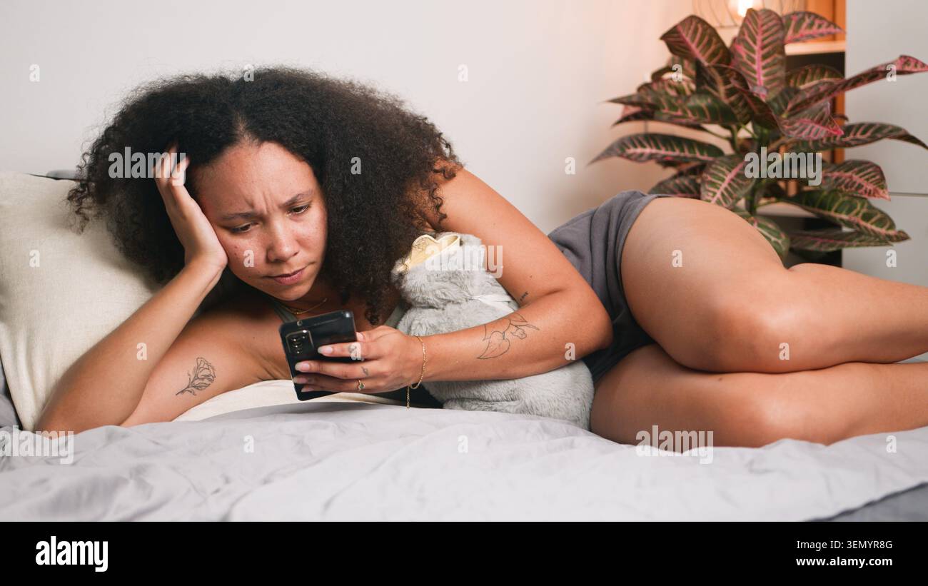 Jeune femme allongée sur le lit regardant smartphone tout en embrassant un animal en peluche Banque D'Images