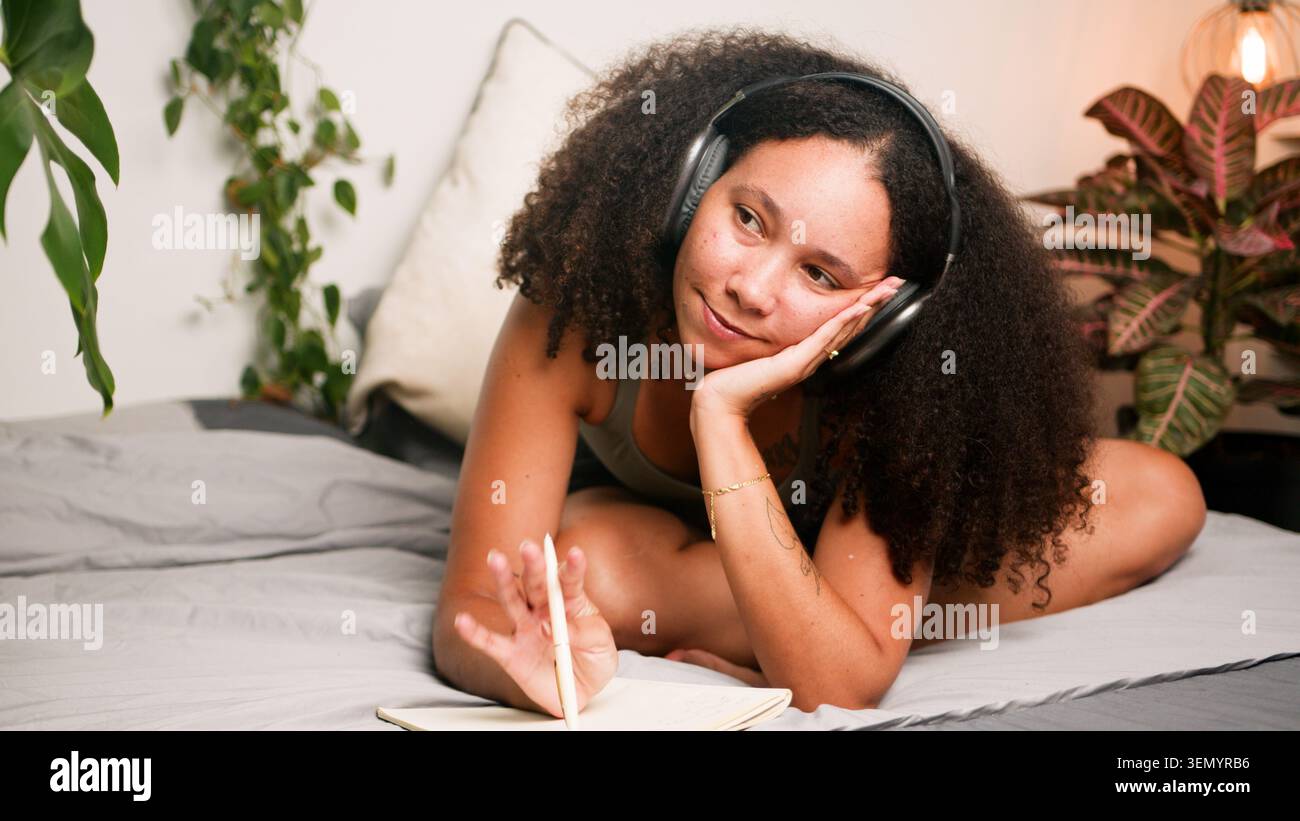 Femme noire se relaxant sur le lit avec des écouteurs journalisant et rêvant dans une chambre confortable Banque D'Images