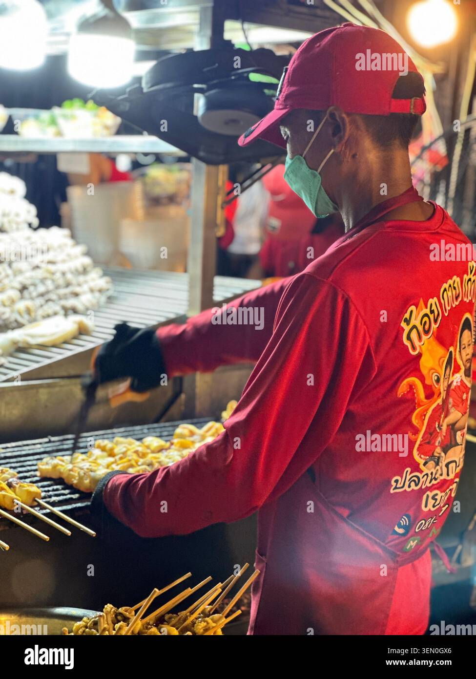 Un cuisinier de rue retourne des rangées de brochettes dorées sur un grill ardent, encadré par de la vapeur, de l’acier et un rouge vif - Image de stock capturée avec un smartphone