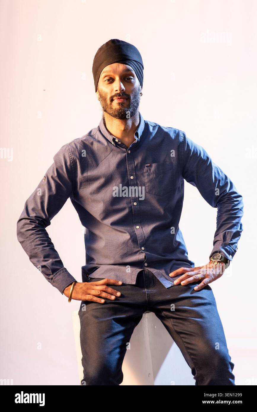 Bigg Taj est assis en toute confiance dans un cadre de studio. Il porte une chemise boutonnée bleue et un Jean. Une toile de fond blanche met en valeur sa pose et son expression pendant t Banque D'Images