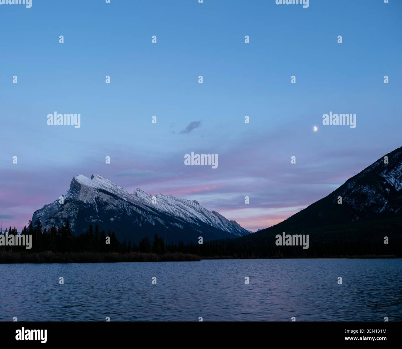 Montagnes enneigées sous un ciel crépusculaire avec un croissant de lune visible au-dessus d'un lac calme. Banque D'Images