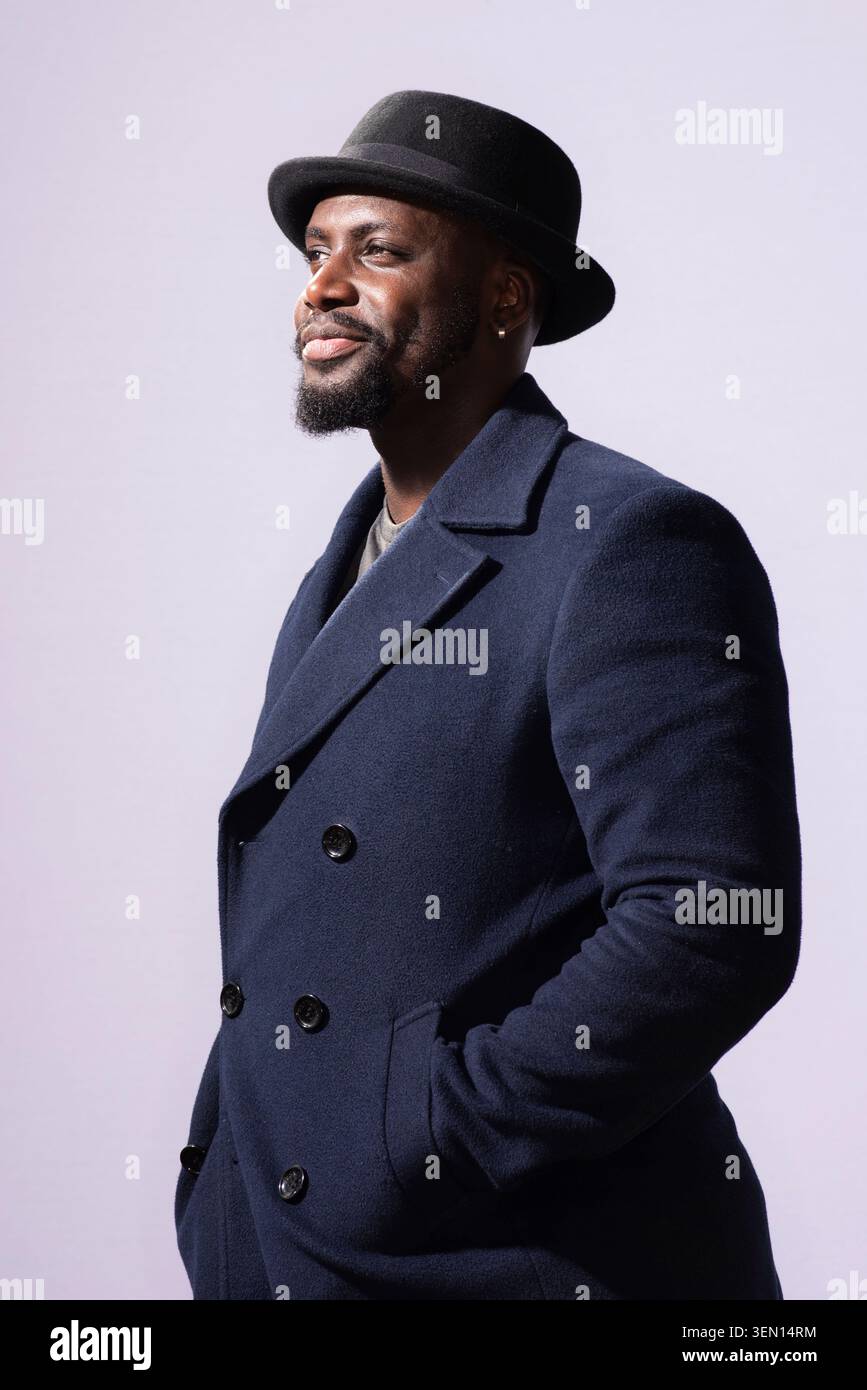 Derek Owusu se tient debout dans un studio portant un manteau foncé et un chapeau noir. Il a un léger sourire et regarde sur le côté, mettant en valeur son style et sa confiance d Banque D'Images