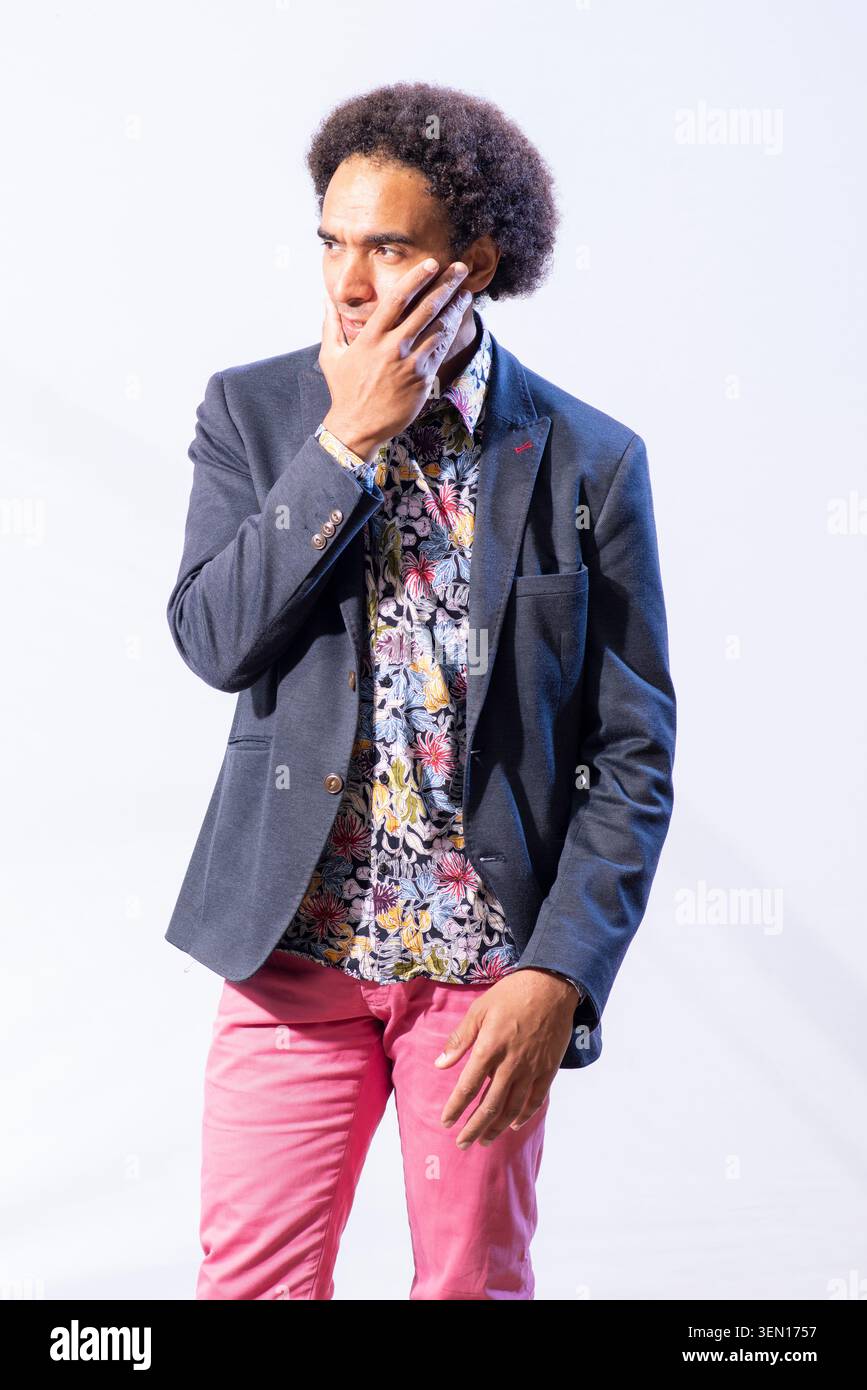 Joseph Coelho se tient debout la main sur le visage, portant une chemise à motifs et un blazer. Il est dans un espace studio lumineux avec un fond Uni. Banque D'Images