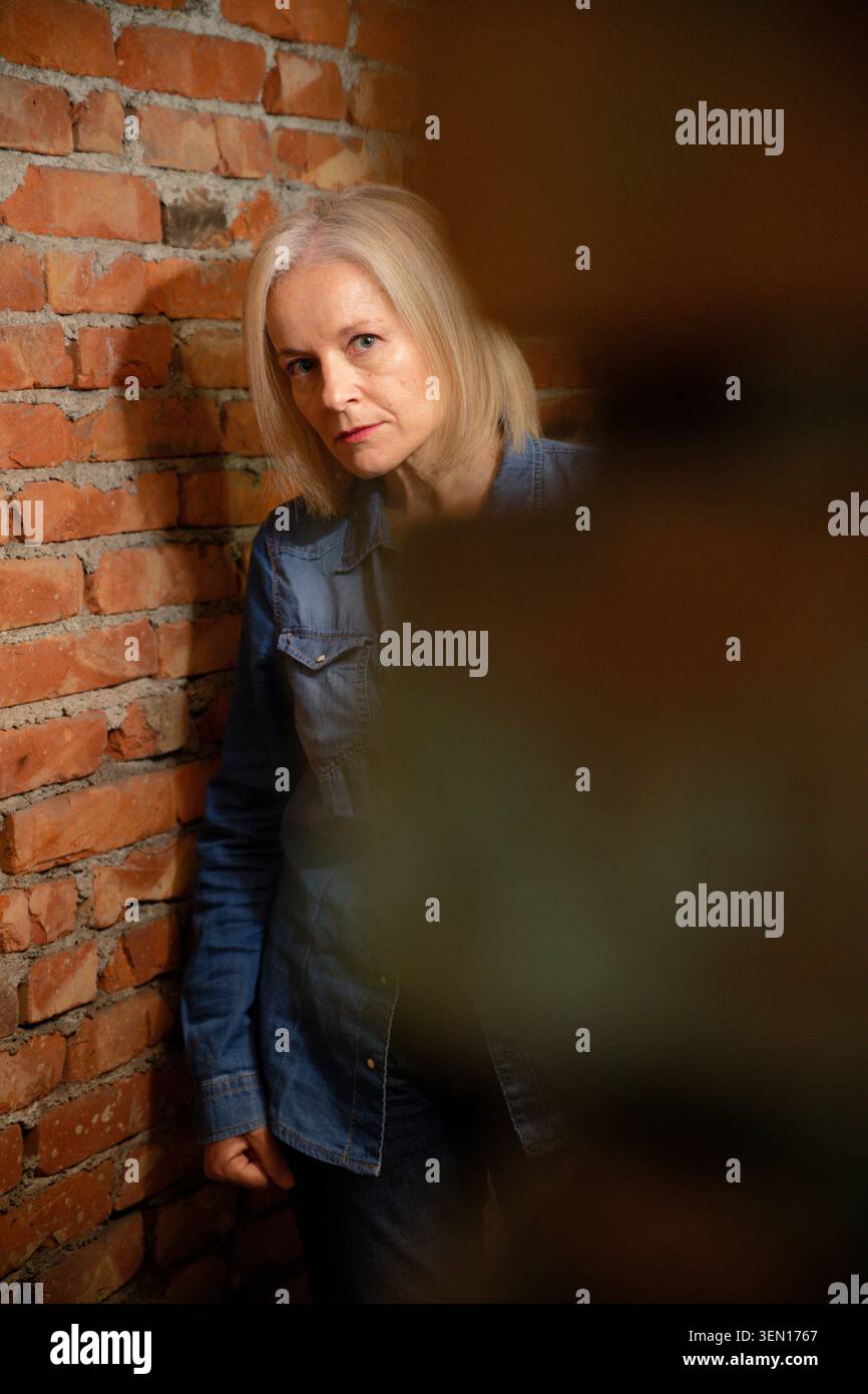 Femme avec de longs cheveux blonds en chemise en denim s'appuie contre le mur de briques exposées. Banque D'Images