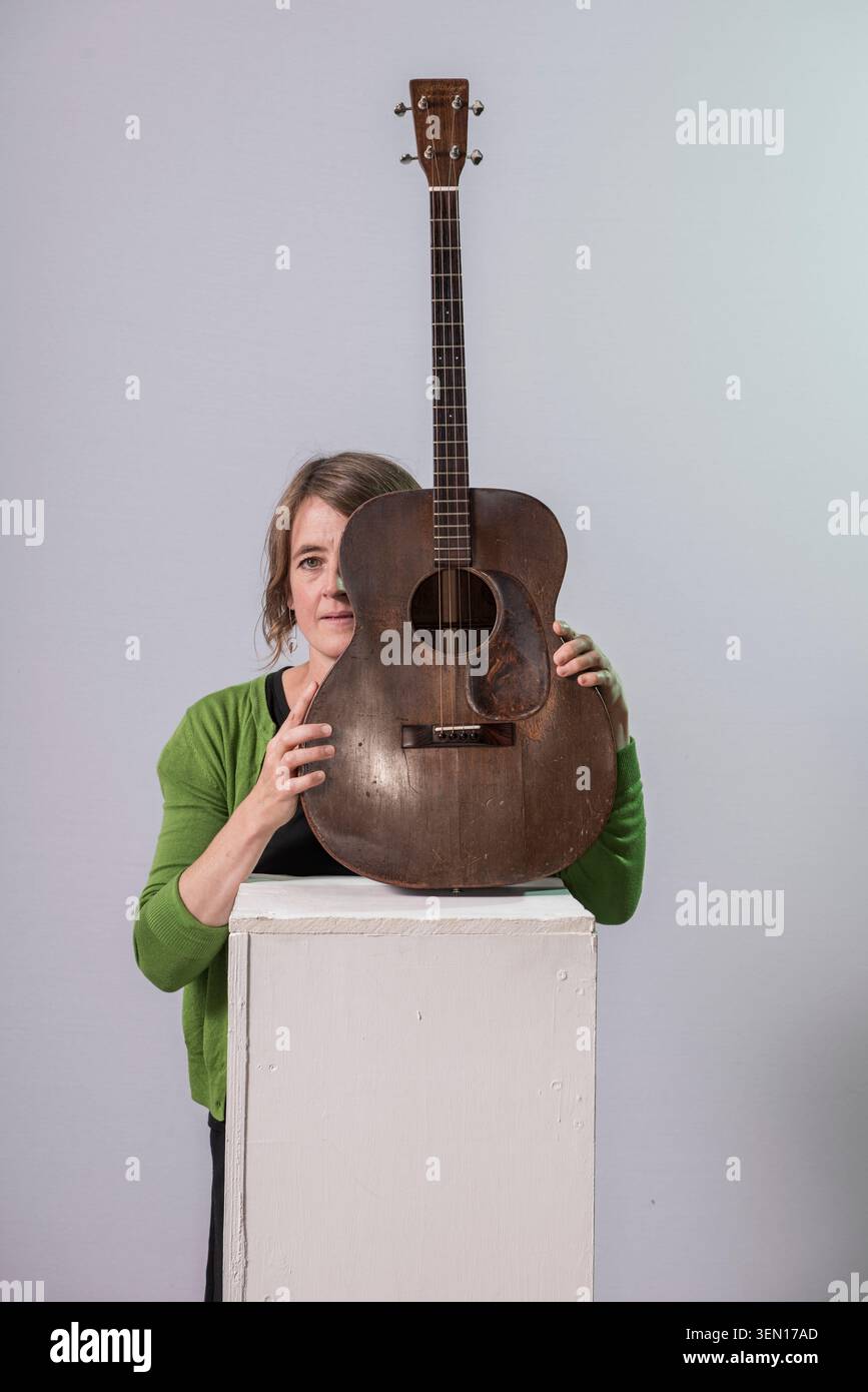 Karine Polwart se tient derrière un podium blanc et tient une vieille guitare. Le fond est clair et l'accent est mis sur elle et la guitare. Elle semble engag Banque D'Images