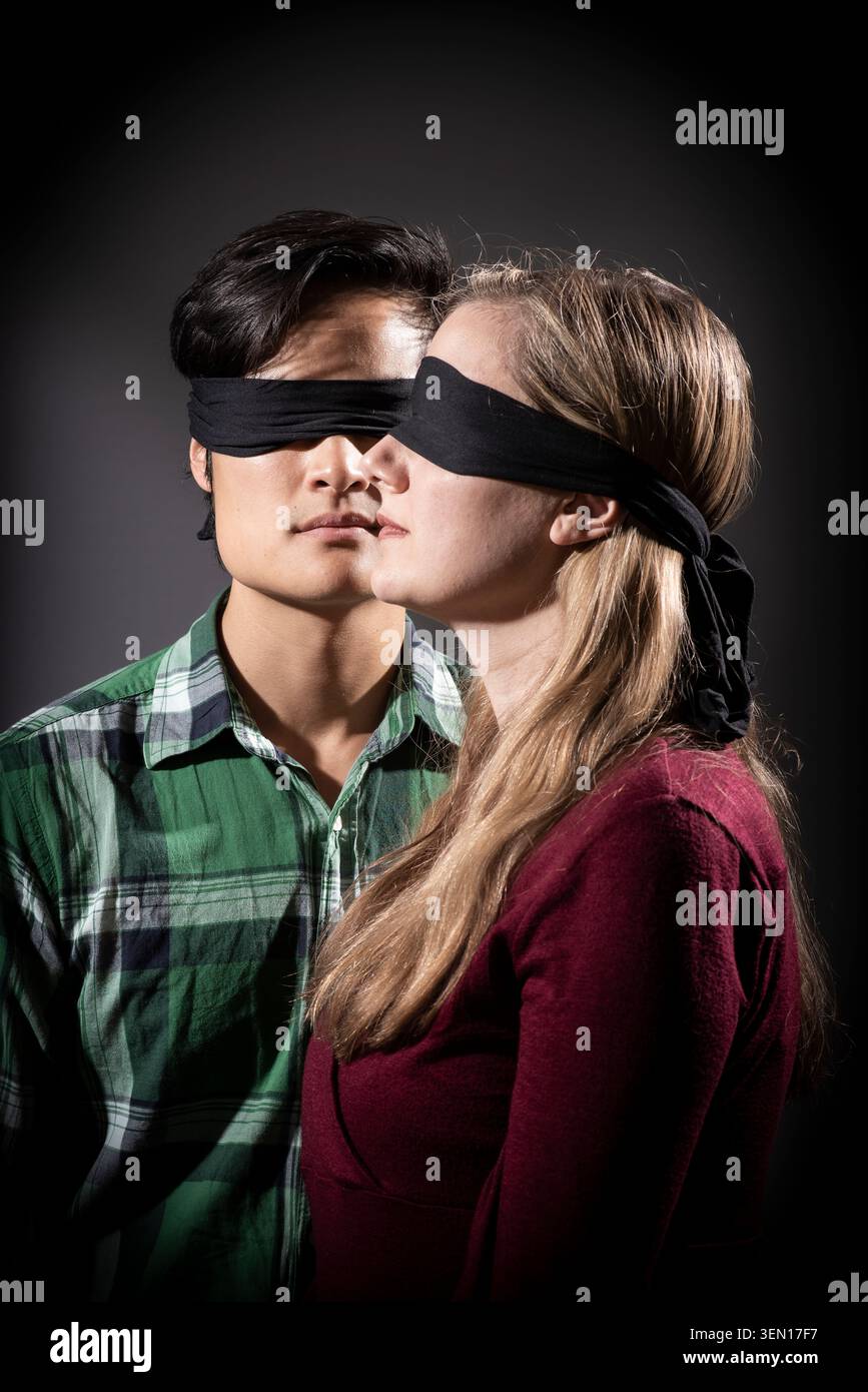 Katie et Kevin participent à une activité les yeux bandés dans un studio. Ils se tiennent proches les uns des autres, portant des bandeaux noirs. L'éclairage ajoute une focalisation Banque D'Images
