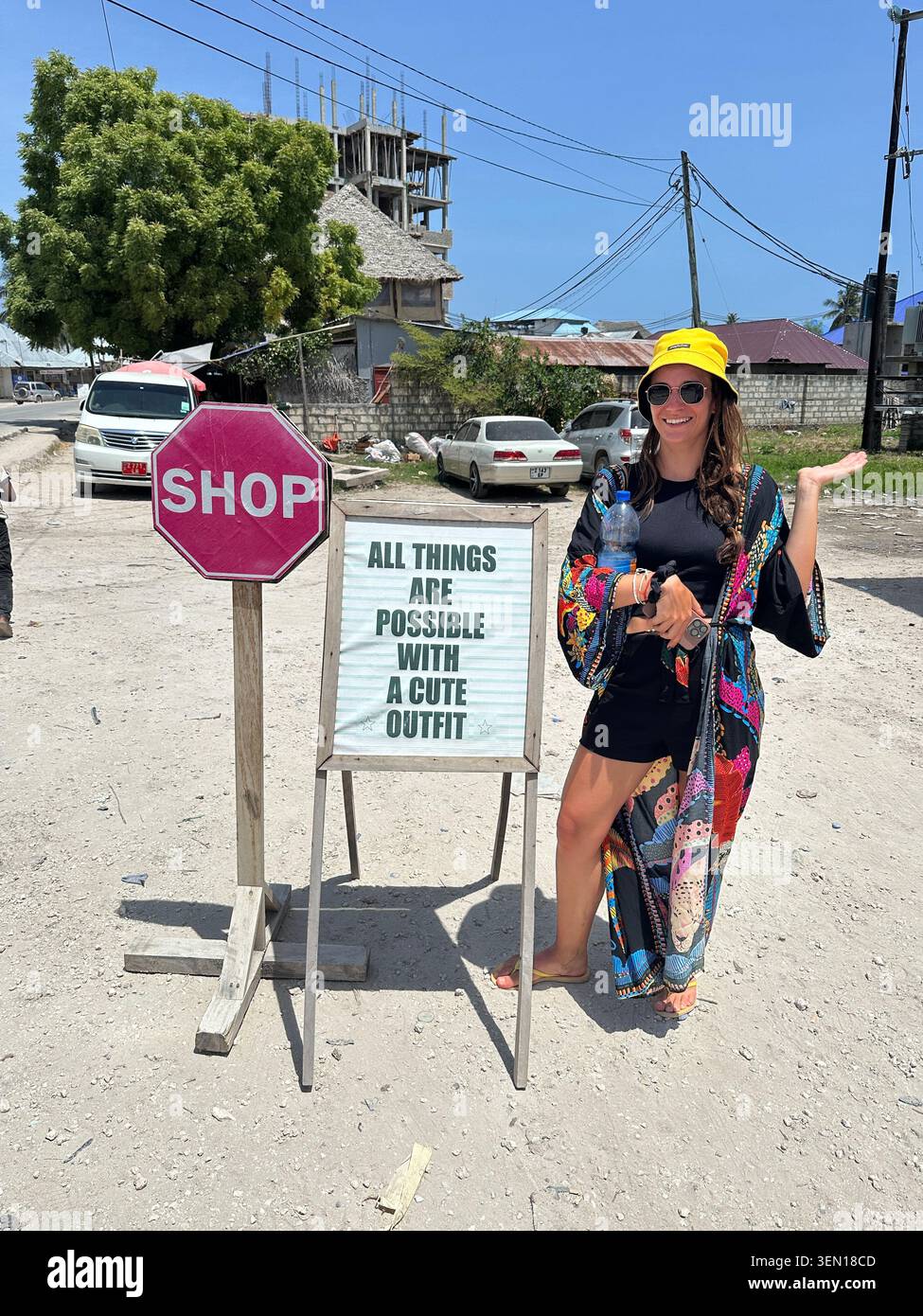 Un voyageur pose à côté d'un panneau humoristique indiquant « Magasin : Tout est possible avec une tenue mignonne » sur une rue insulaire ensoleillée. - Image de stock capturée avec un smartphone