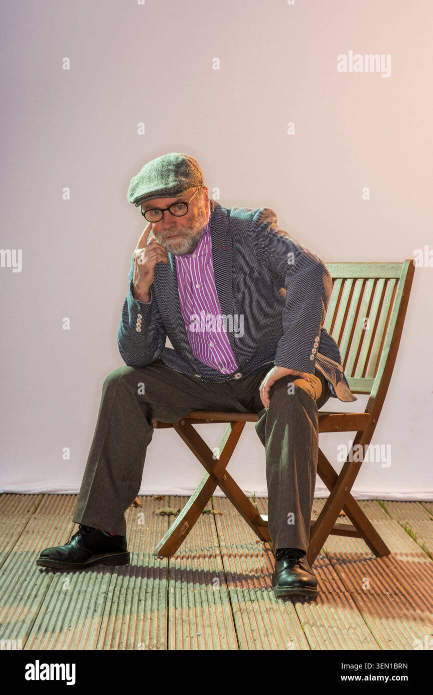 Malachi O'Doherty est assis sur une chaise en bois, portant des lunettes et une casquette. Il pose soigneusement, avec une main sur son menton, dans un cadre de studio avec l doux Banque D'Images