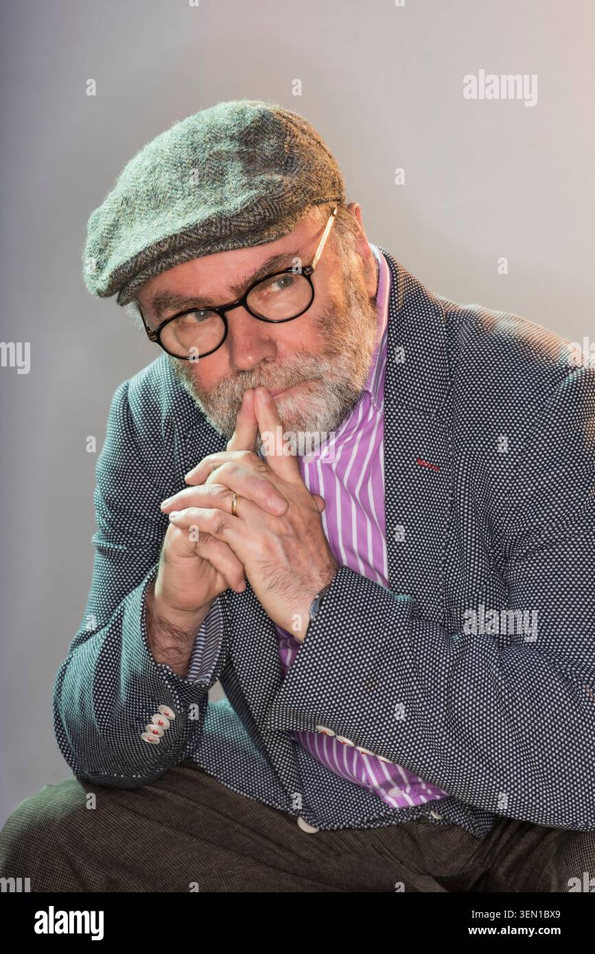 Malachi O'Doherty est assis dans un studio avec une expression réfléchie. Il porte une casquette et des lunettes. Le réglage est simple avec un éclairage doux. Banque D'Images