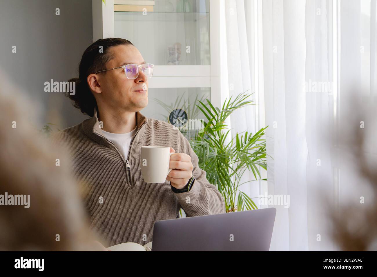 Homme d'âge moyen profitant d'une pause café tout en travaillant à distance près de la fenêtre lumineuse. Pause paisible, équilibre entre vie professionnelle et vie privée, productivité et bureau à domicile calme Banque D'Images
