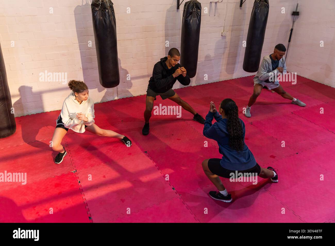 Groupe diversifié faisant des fentes latérales dans la salle de boxe, tapis rouges, sacs lourds avec adolescent, portant des vêtements de sport Banque D'Images