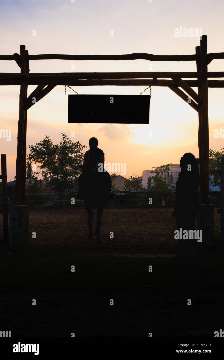 Balikpapan, Indonésie - 1er mai 2026. Une silhouette spectaculaire d'un cheval et cavalier encadrée par une passerelle en bois rustique au coucher du soleil. Banque D'Images