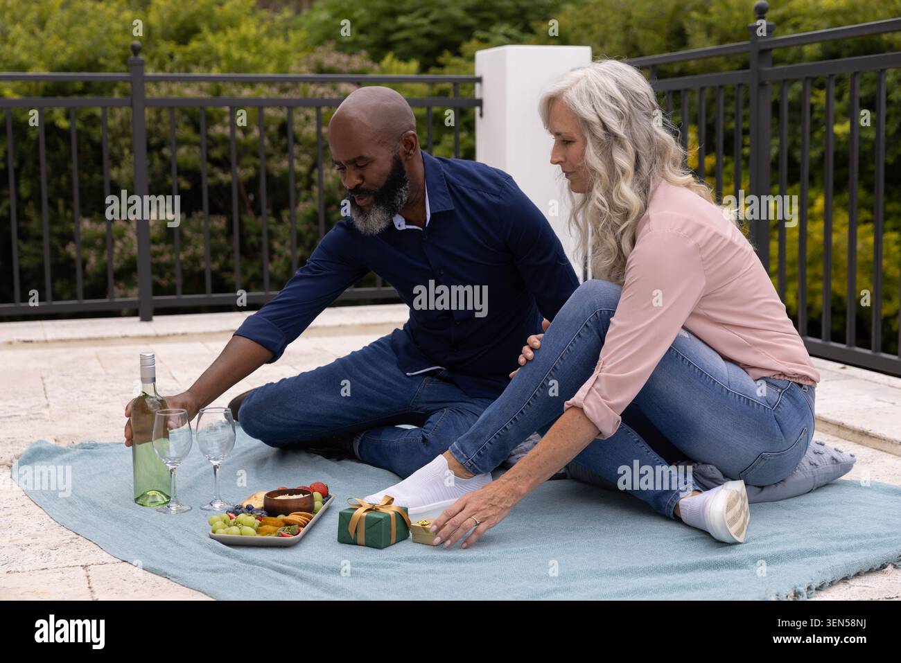 Couple diversifié assis sur une couverture de pique-nique bleu clair sur un patio en pierre, partageant vin blanc et plat Banque D'Images