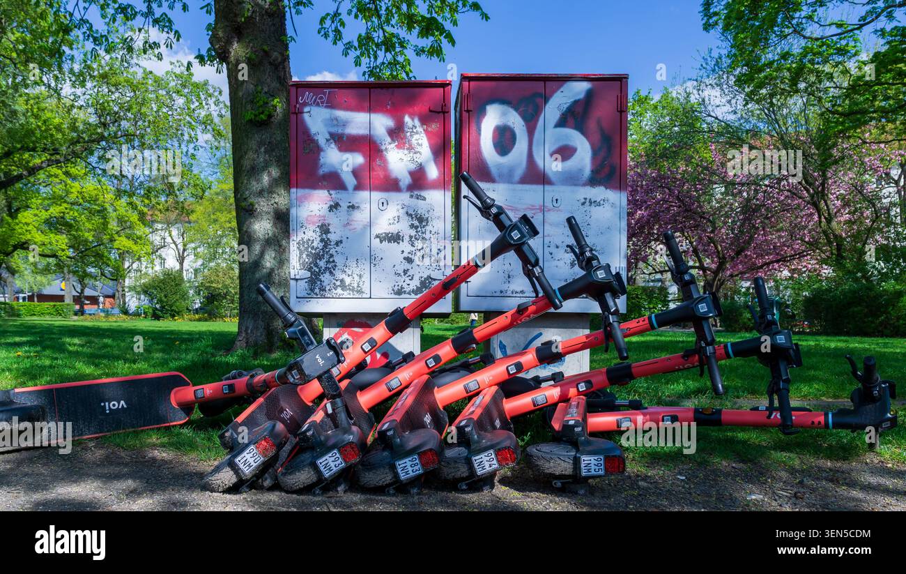Plusieurs scooters électriques partagés sont empilés sur le sol devant des boîtes utilitaires couvertes de graffiti dans un parc public, e-scooter, e-scooter rouge. Banque D'Images