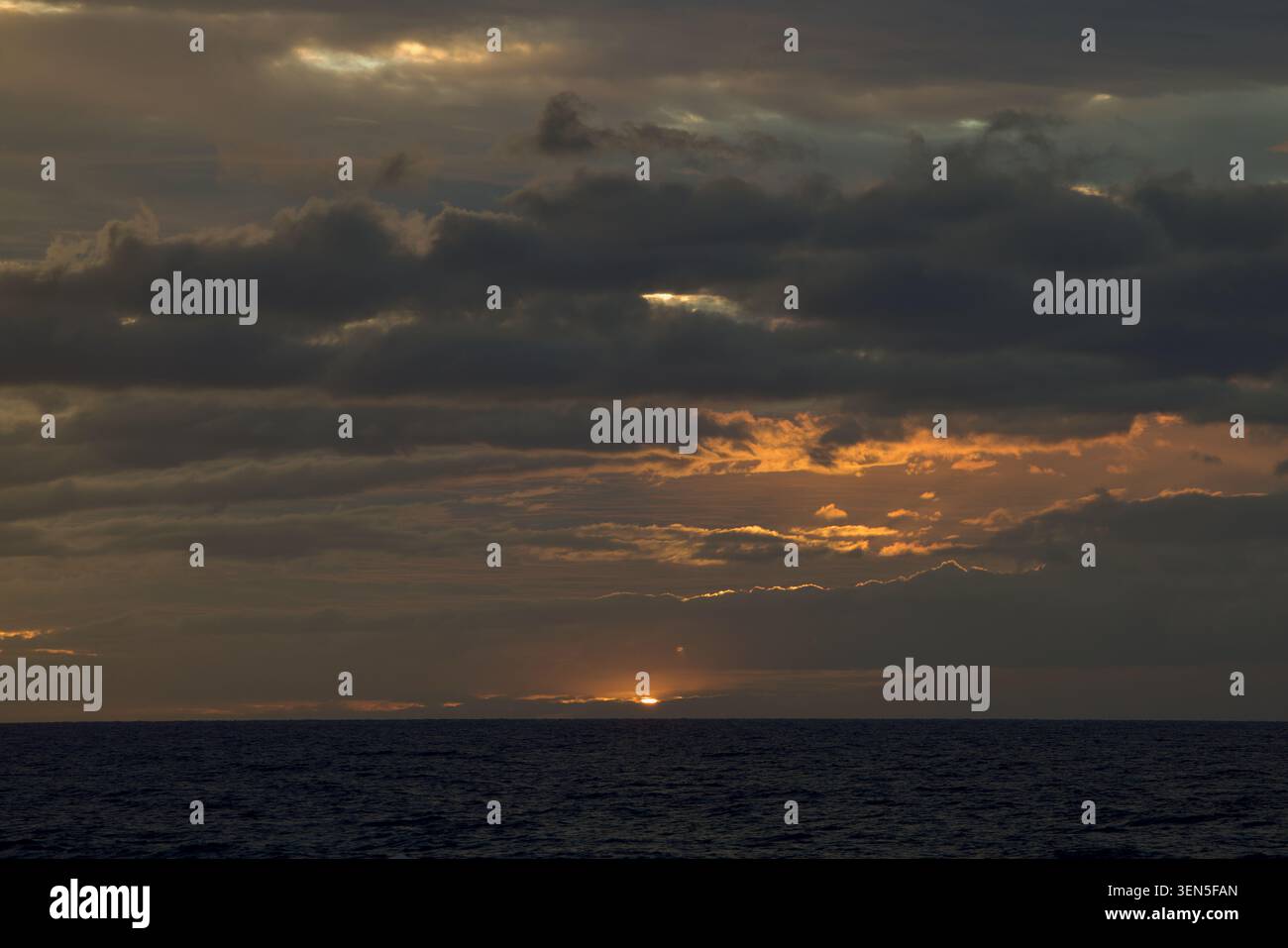 Coucher de soleil Moody au crépuscule sur un vaste océan ouvert sombre. De lourds nuages de tempête gris couvrent le ciel, de minces filtres de lumière ambre chaude et dorée pâle à travers les espaces nuageux. Banque D'Images