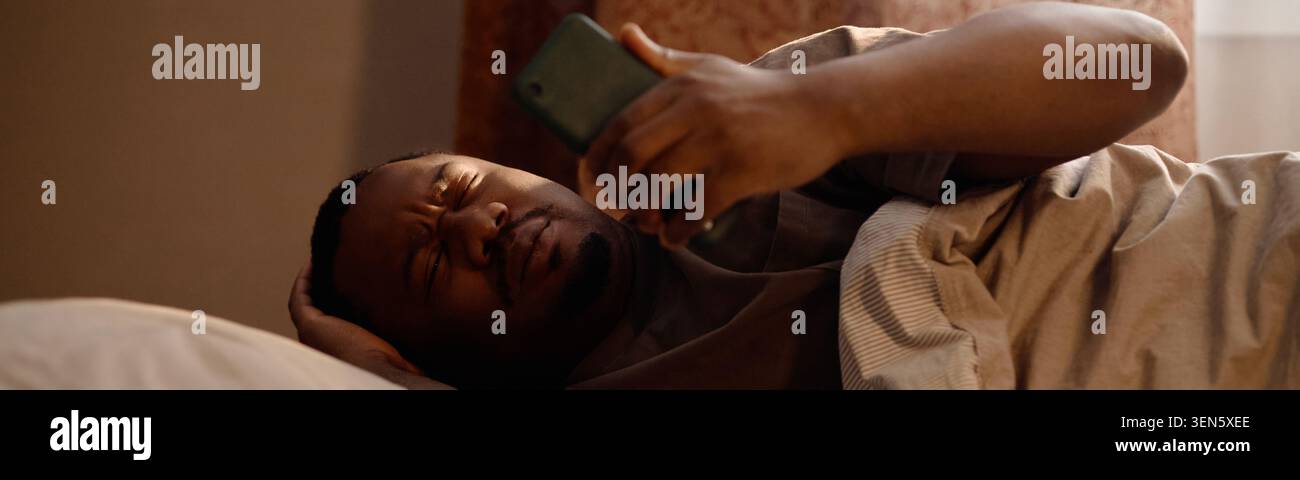 Homme noir utilisant un smartphone dans le lit montrant la fatigue numérique à la maison Banque D'Images