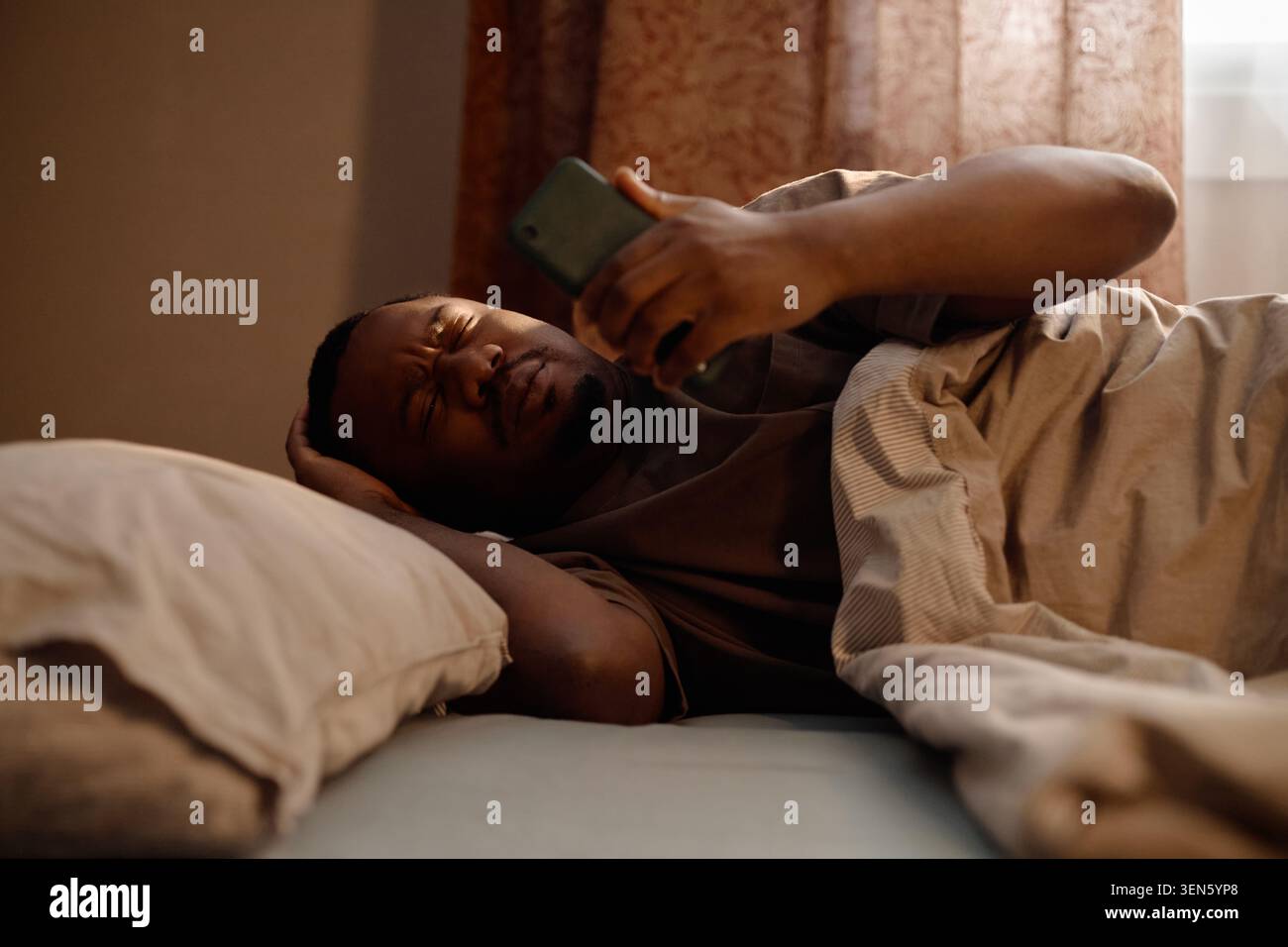 Homme noir vérifiant smartphone dans le lit avec la tension oculaire à la maison Banque D'Images