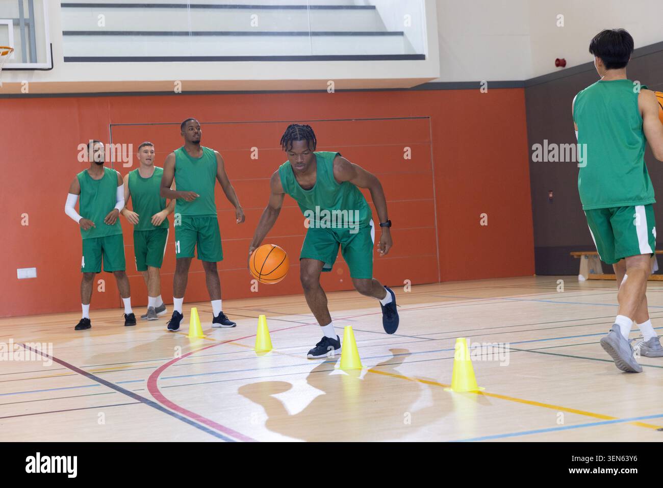 Divers coéquipiers masculins pratiquant l'exercice dribble dans le gymnase portant un équipement vert avec une balle orange, des cônes Banque D'Images