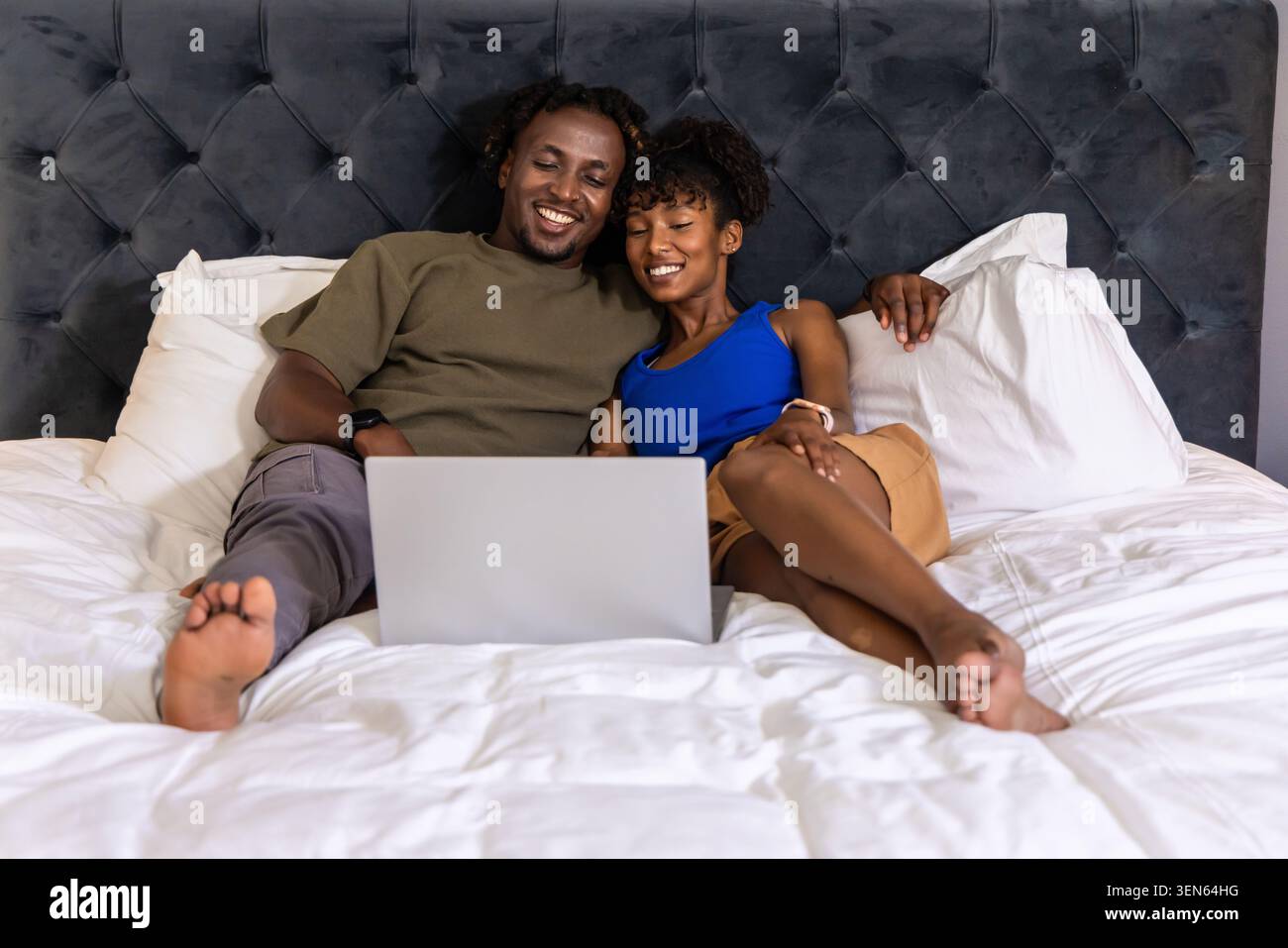 Couple afro-américain incliné dans le lit dans la chambre, portant un tee-shirt olive et un réservoir bleu, à l'aide d'un ordinateur portable Banque D'Images