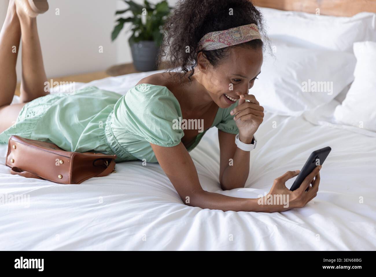 smartphone brillant sur le lit blanc, sac à main marron reposant à côté de l'oreiller contre la tête de lit avec plante Banque D'Images