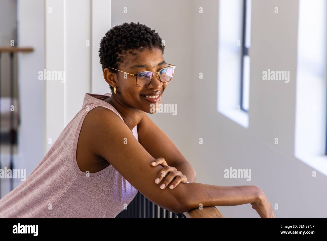 Femme africaine appuyée sur une balustrade, souriant dans le hall avec des fenêtres, portant des lunettes en or, top rose Banque D'Images
