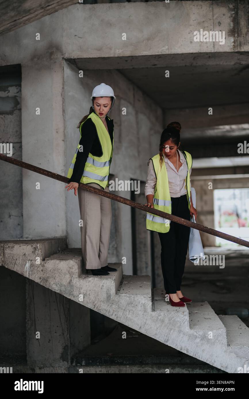 Deux femmes ingénieurs inspectant une cage d'escalier de chantier inachevée en gilets de sécurité et casque de sécurité Banque D'Images