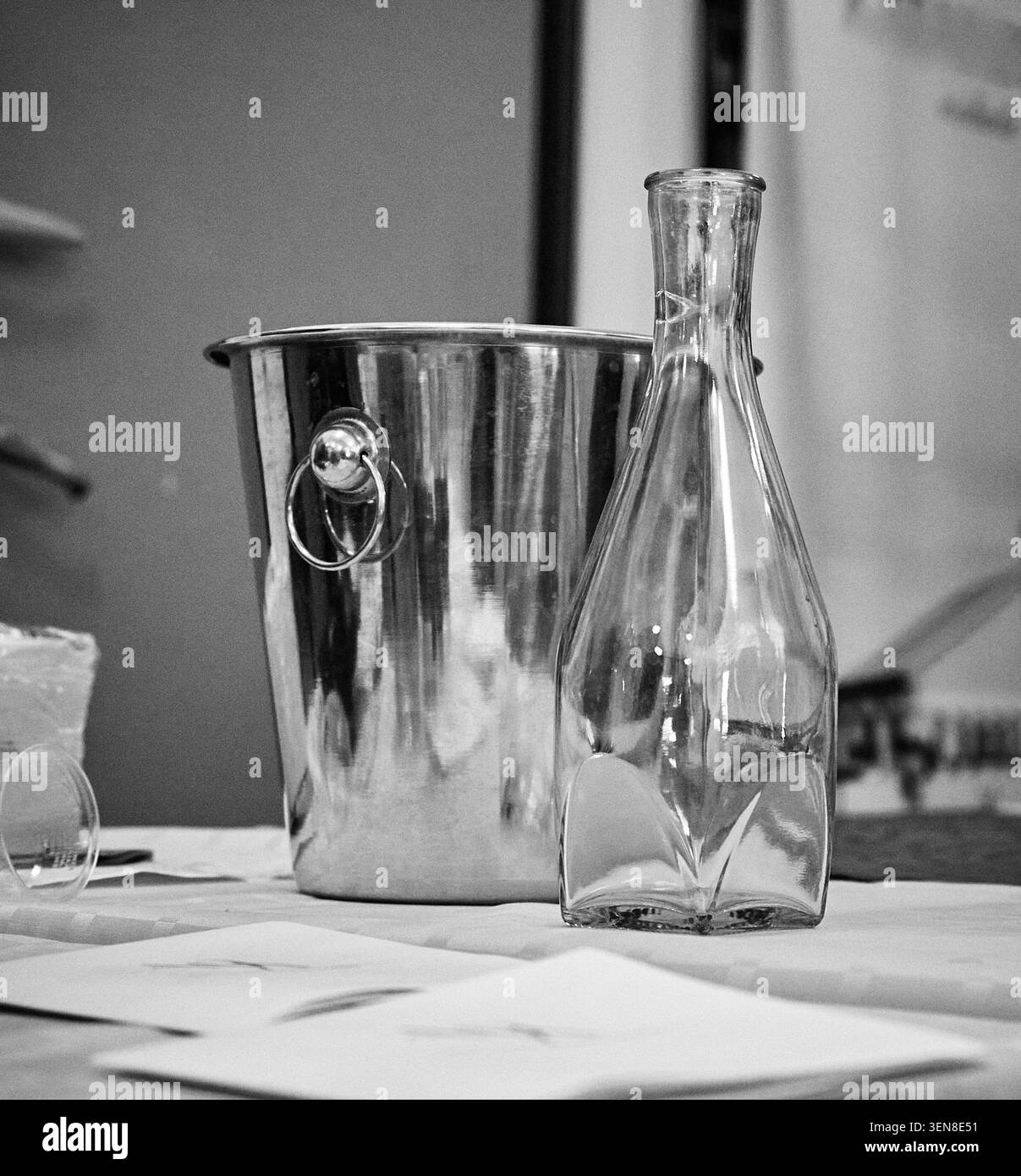 Bouteille en verre et seau à glace en acier inoxydable sont positionnés sur une table avec une nappe blanche, accompagnés de papiers éparpillés en arrière-plan Banque D'Images