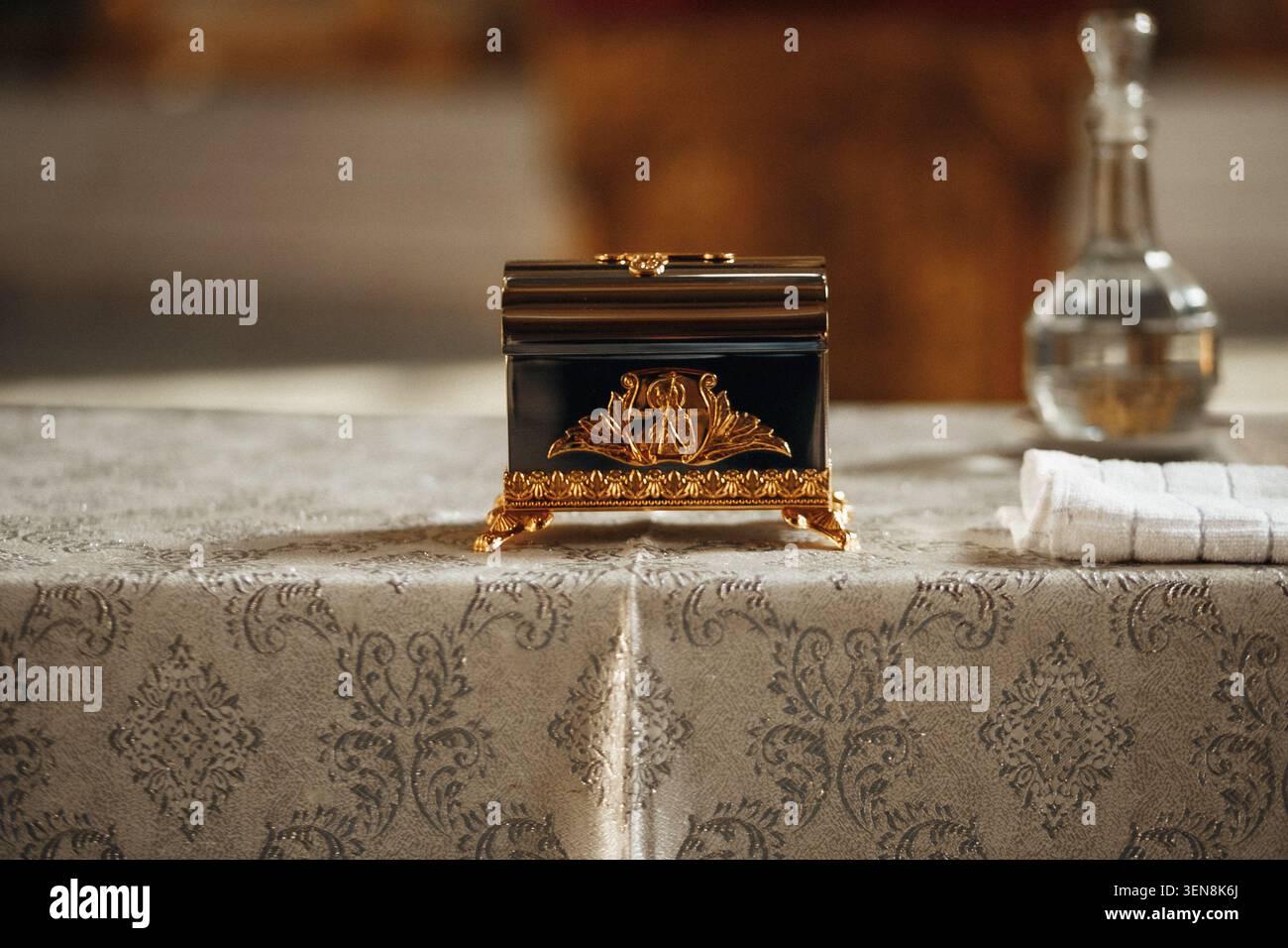 Élégante boîte noire avec couvercle orné doré repose sur une nappe damassée. Carafe en verre et lin plié se trouvent à proximité, ajoutant une touche raffinée. Flou de mise au point Banque D'Images