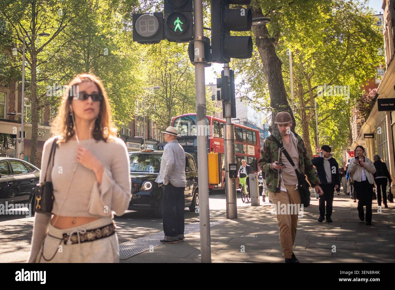 LONDRES - 23 AVRIL 2026 : Kensington High Street – jeune femme avec des lunettes de soleil dans une rue ensoleillée animée de Londres avec des bus rouges Banque D'Images