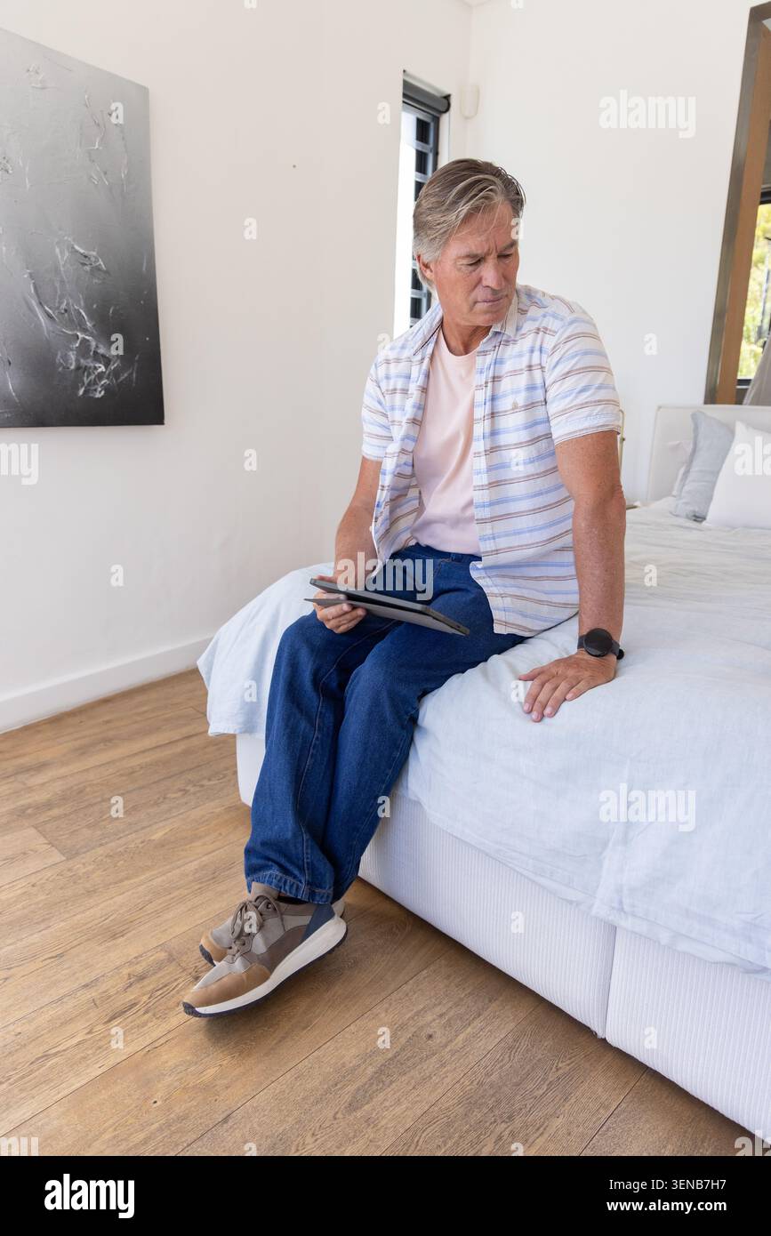 Homme mûr assis sur le bord du lit tenant la tablette, portant une chemise rayée, un Jean bleu, des baskets tan Banque D'Images