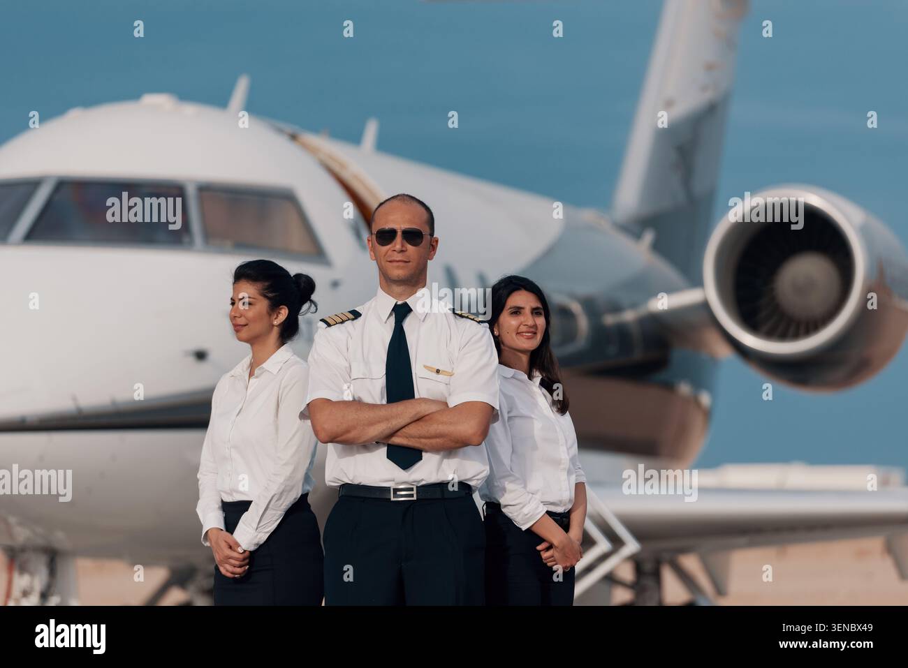 Pilote de jet privé confiant en lunettes de soleil aviateur avec les bras croisés debout avec deux hôtesses de bord féminines sur le tarmac de l'aéroport avant le luxe Banque D'Images