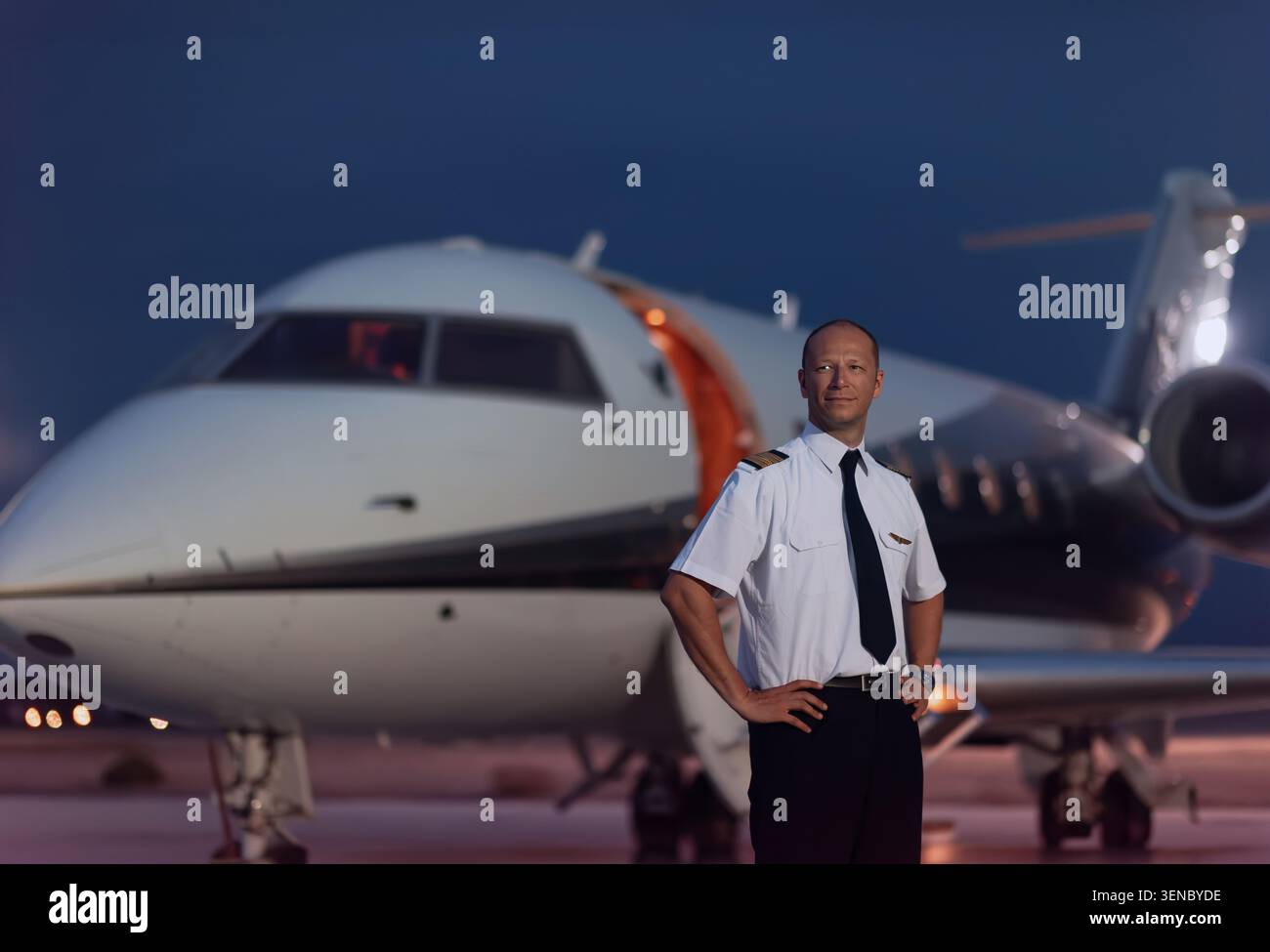 Pilote de ligne masculin confiant en uniforme debout avec les mains sur les hanches sur le tarmac de l'aéroport à côté des jets privés de luxe au crépuscule idéal pour la carrière d'aviation et Banque D'Images