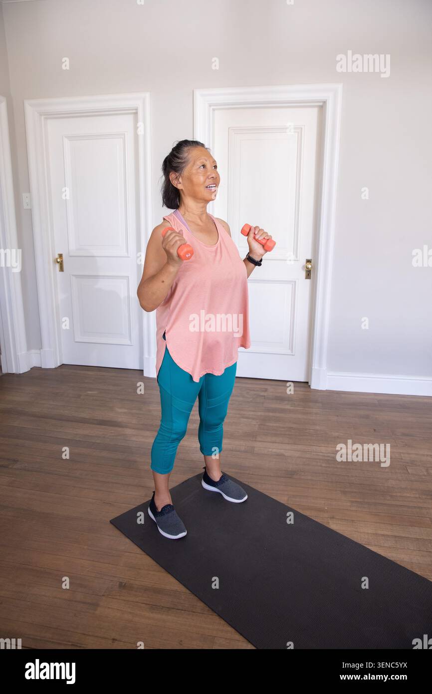 Femme senior soulevant des haltères de corail sur tapis à la maison, portant un haut de corail, leggings sarcelle, smartwatch Banque D'Images