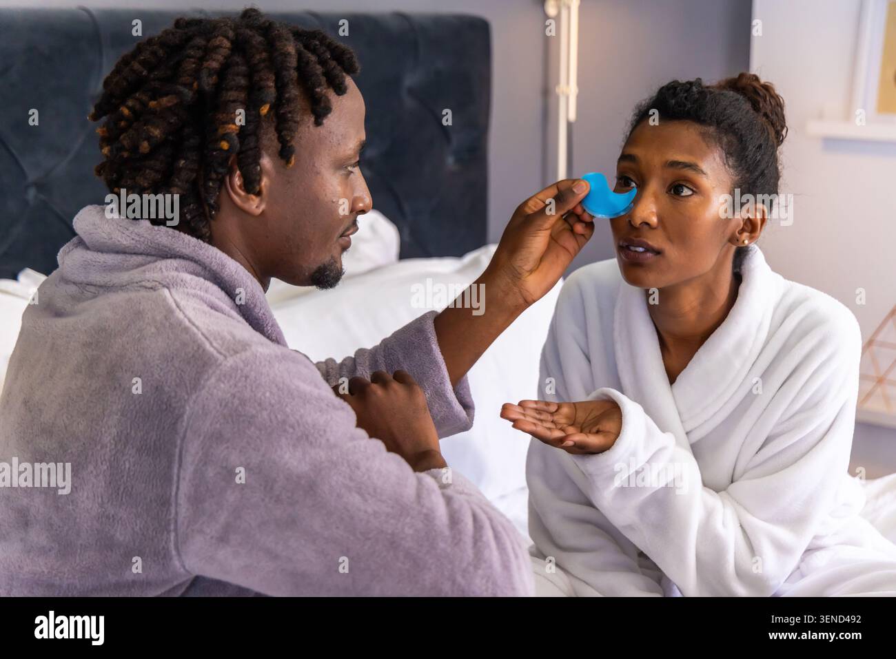 Couple assis dans la chambre sur le lit portant des robes, homme appliquant un dispositif facial bleu près de l'oeil de la femme Banque D'Images