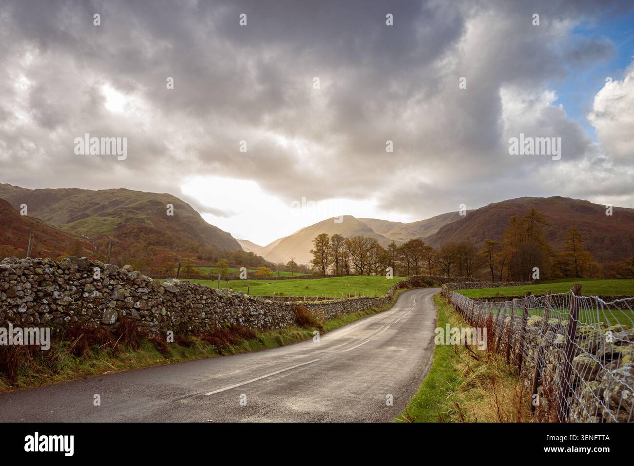 Une route rurale tranquille serpente entre de vieux murs de pierre à travers une vallée verdoyante dans le Lake District, Cumbria, avec des nuages spectaculaires et orageux qui se rassemblent au-dessus. Banque D'Images