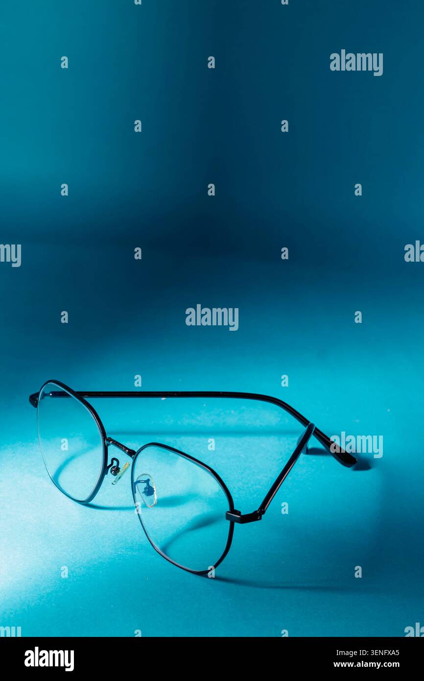 Une paire de lunettes rondes élégantes en métal noir bordées de fils assis sur une surface bleue éclatante sous un éclairage ponctuel spectaculaire, représentant la vision, la clarté, l'éducation et l'intelligence. Banque D'Images