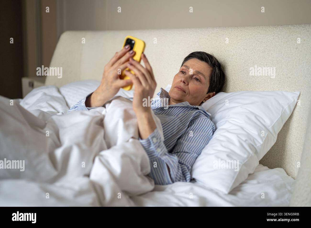 Femme senior détendue faisant défiler les flux multimédia en ligne sur smartphone tout en étant couché dans le lit dans la chambre. Banque D'Images