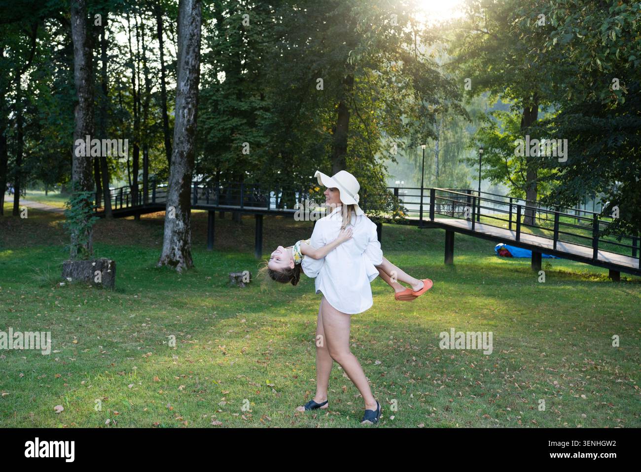 Une femme portant un chapeau de soleil et une chemise surdimensionnée balançant ludique son enfant dans un parc verdoyant. La scène est baignée de soleil chaud en fin d'après-midi Banque D'Images