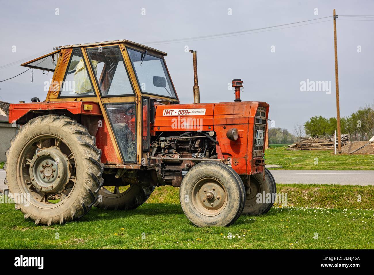 Vinkovci, Croatie - avril 10.2026 :Un tracteur rouge vintage garé sur un champ herbeux à côté d'une ferme rurale, montrant des pneus usés, moteur exposé et m Banque D'Images